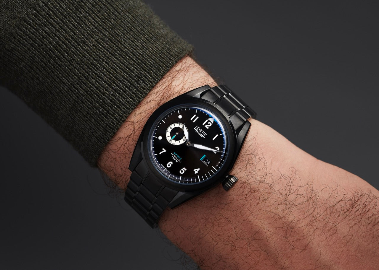 Black watch with a black dial, white numerals and blue colour accents. Includes a seconds hand dial and date display. Shown on a person's wrist.