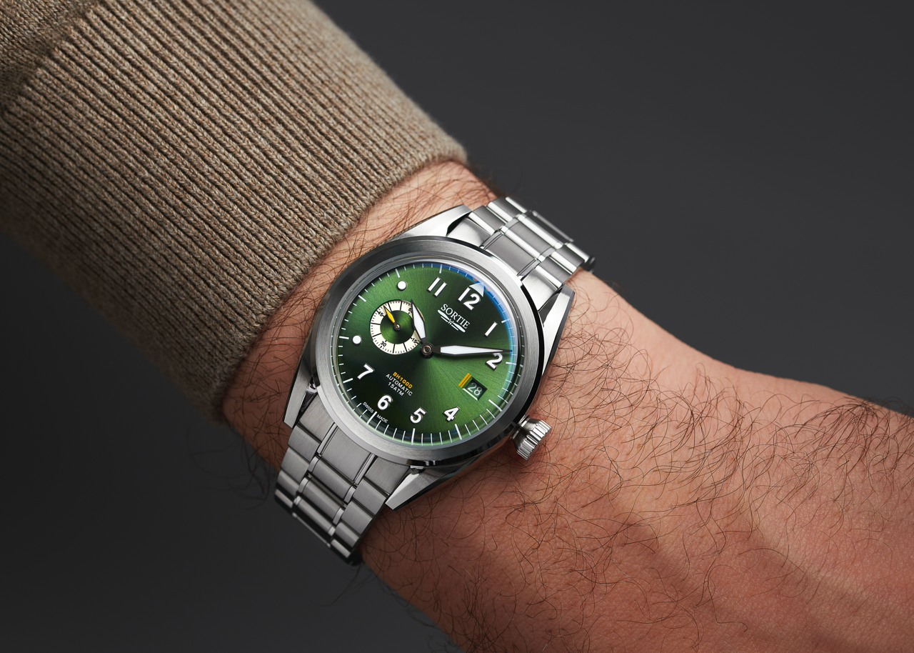 Silver stainless steel watch with a green dial, white numerals and yellow colour accents. Includes a seconds hand dial and date display. Shown on a person's wrist.